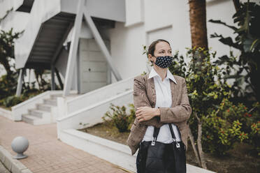 Businesswoman wearing protective face mask standing with arms crossed against building - DMGF00494