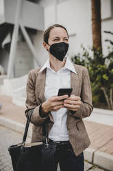 Geschäftsfrau mit Schutzmaske, die ein Smartphone in der Hand hält und an einem Gebäude steht, während sie wegschaut - DMGF00497