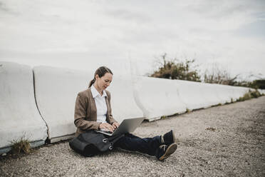 Unternehmerin arbeitet am Laptop, während sie an der Leitplanke am Straßenrand sitzt - DMGF00510