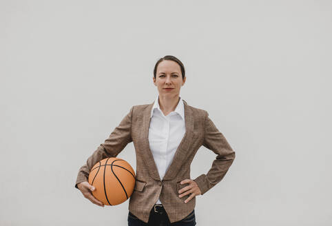 Selbstbewusste weibliche Fachkraft mit Basketball, die mit der Hand auf der Hüfte vor einer weißen Wand steht - DMGF00511