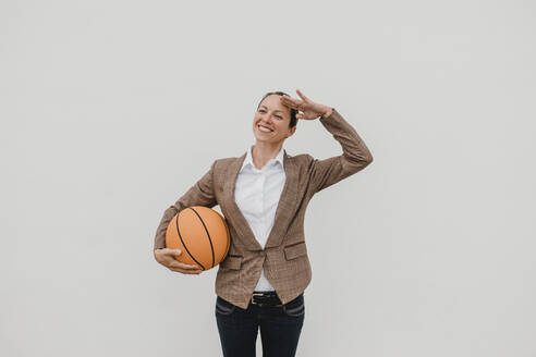 Lächelnde weibliche Fachkraft mit Basketball, die vor einer weißen Wand steht und grüßt - DMGF00518