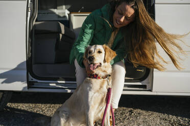 Mid adult woman stroking dog sitting at camper van during sunny day - MPPF01538