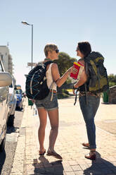 Smiling female friends with backpack talking on footpath during sunny day - AJOF01149