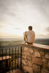 Junger Mann sitzt bei Sonnenuntergang auf einer Stützmauer - ACPF01170