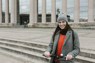 Smiling woman with electric push scooter standing near building - XLGF01241