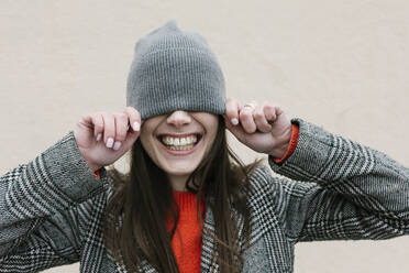 Smiling woman covering eyes through knit hat against wall - XLGF01244