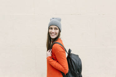 Smiling young woman with backpack looking away in front of wall - XLGF01246