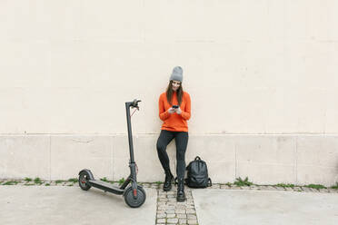 Woman using smart phone by electric push scooter while leaning on wall - XLGF01250