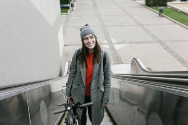 Nachdenkliche Frau, die einen Elektroroller hält, während sie auf einer Rolltreppe in der Stadt steht - XLGF01256