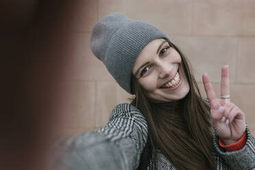 Cheerful young woman in warm clothing showing peace sign against wall - XLGF01264