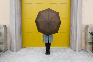 Woman holding umbrella while standing against yellow door - XLGF01273