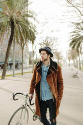 Hipster-Mann mit Hut steht mit Fahrrad in der Stadt und schaut weg - GMCF00023