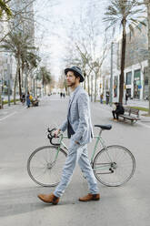 Männlicher Berufstätiger mit Hut beim Spaziergang mit dem Fahrrad in der Stadt - GMCF00033