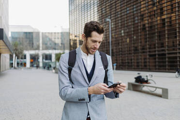 Businessman with backpack using smart phone in city - GMCF00037