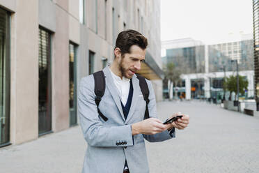 Männlicher Unternehmer mit Rucksack, der ein Mobiltelefon benutzt, während er an einem Gebäude steht - GMCF00038