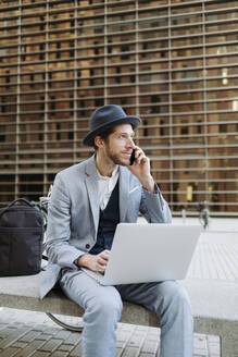 Männlicher Unternehmer mit Hut sitzt mit Laptop auf einer Bank und spricht mit seinem Smartphone - GMCF00045