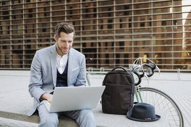 Männlicher Berufstätiger, der auf einer Bank sitzend am Laptop arbeitet - GMCF00046