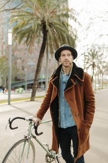 Male hipster standing with bicycle looking away - GMCF00063