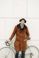 Happy man in coat standing with bicycle against wall - GMCF00067