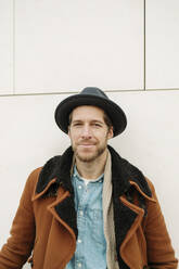 Male hipster wearing coat and hat against white wall - GMCF00069