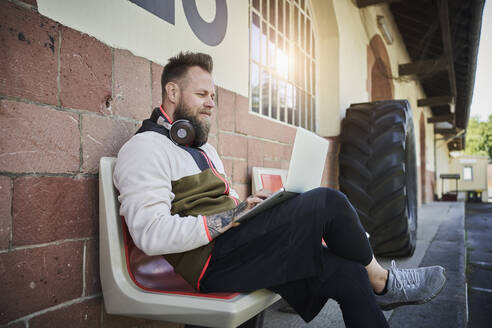 Bärtiger männlicher Fitnesstrainer, der einen Laptop benutzt, während er auf einer Bank vor einem Fitnessstudio sitzt - RORF02607