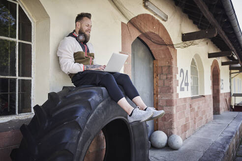Männlicher Fitnesstrainer, der einen Laptop benutzt, während er auf einem Reifen vor einem Fitnessstudio sitzt - RORF02608