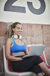 Weiblicher Fitnesstrainer mit Laptop, während er auf einer Bank an der Wand sitzt - RORF02614