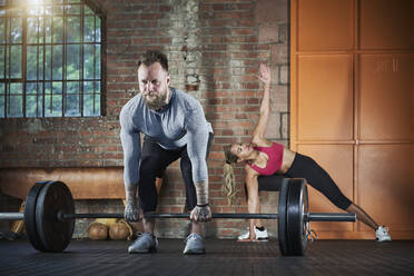 Sportsman lifting barbell while female athlete exercising in background at gym - RORF02658