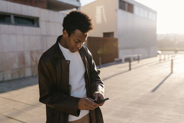 Mid adult man text messaging through smart phone while standing on footpath during sunset - TCEF01619