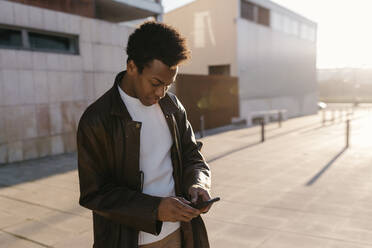 Mittelgroßer erwachsener Mann, der während des Sonnenuntergangs auf dem Fußweg steht und mit seinem Smartphone SMS schreibt - TCEF01619
