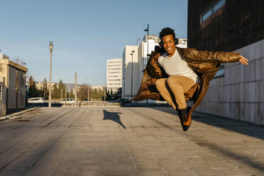 Smiling man wearing headphones jumping while practicing dance during sunset - TCEF01630