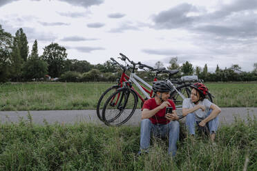 Father holding smart phone while sitting with daughter talking at agricultural field - OGF00921