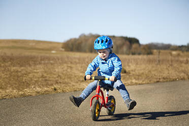 Verspielter Junge sitzt mit gespreizten Beinen auf einem Balance-Bike auf der Straße - SEBF00293