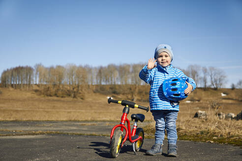 Niedlicher Junge mit Fahrradhelm, der mit der Hand winkt, während er neben einem Gleichgewichtsfahrrad auf der Straße steht - SEBF00296