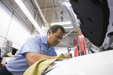 Ein hispanischer Mechaniker lehnt sich in den Motor eines Autos, an dem er in einer Autowerkstatt arbeitet - MINF15829