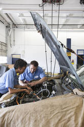 Männliche und weibliche Mechaniker unterhalten sich, während sie sich den Motor in einer Autowerkstatt ansehen - MINF15856