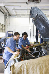 Männliche und weibliche Mechaniker unterhalten sich, während sie sich den Motor in einer Autowerkstatt ansehen - MINF15857