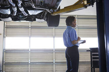 Weiblicher Mechaniker, der ein digitales Tablet hält, das unter einem Autochassis in einer Autowerkstatt steht - MINF15880
