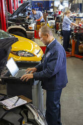 Pacific Islander mechanic typing on a laptop while co-workers work on cars in background - MINF15897