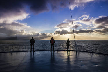 Drei Personen genießen den Sonnenuntergang auf dem Bootsdeck, während das Schiff nach Island fährt - CAVF93612
