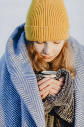 Porträt eines hübschen Mädchens im Teenageralter mit Strickmütze, das sich mit einer Wolldecke und einer Tasse heißem Tee wärmt - OJF00436