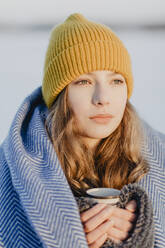 Porträt eines hübschen Mädchens im Teenageralter mit Strickmütze, das sich mit einer Wolldecke und einer Tasse heißem Tee wärmt - OJF00437