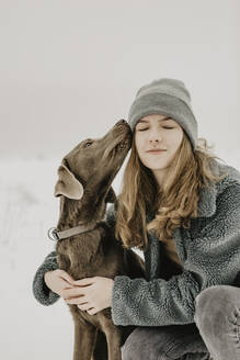 Porträt eines Mädchens im Teenageralter, das im Schnee hockt und einen Labrador Retriever umarmt - OJF00440