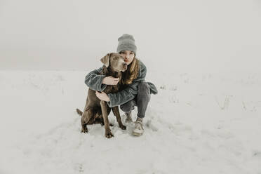 Porträt eines Mädchens im Teenageralter, das im Schnee hockt und einen Labrador Retriever umarmt - OJF00443