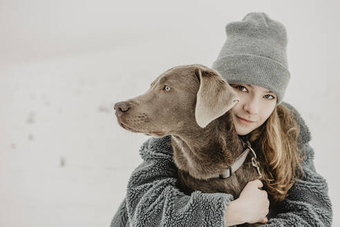 Porträt eines Mädchens im Teenageralter, das im Schnee hockt und einen Labrador Retriever umarmt - OJF00446