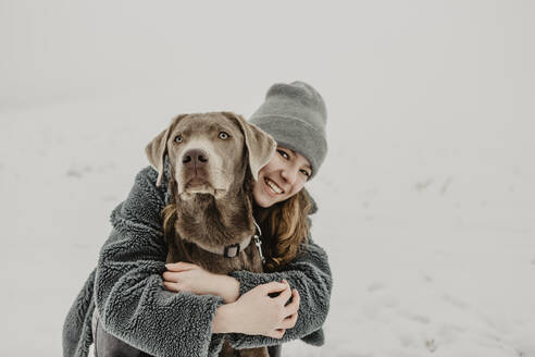 Porträt eines Mädchens im Teenageralter, das im Schnee hockt und einen Labrador Retriever umarmt - OJF00448