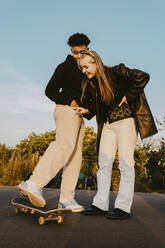 Männliche und weibliche Freunde stehen bei Sonnenuntergang im Park neben einem Skateboard - MASF22074