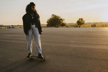 Woman looking away while standing on skateboard in park during sunset - MASF22077