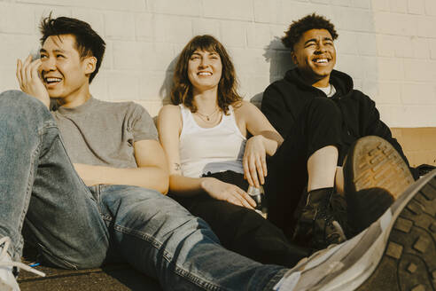 Lächelnde männliche und weibliche Freunde, die an einem sonnigen Tag auf einem Fußweg vor einer Mauer sitzen - MASF22098