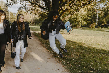 Cheerful woman jumping with skateboard while female friends walking by in park - MASF22116